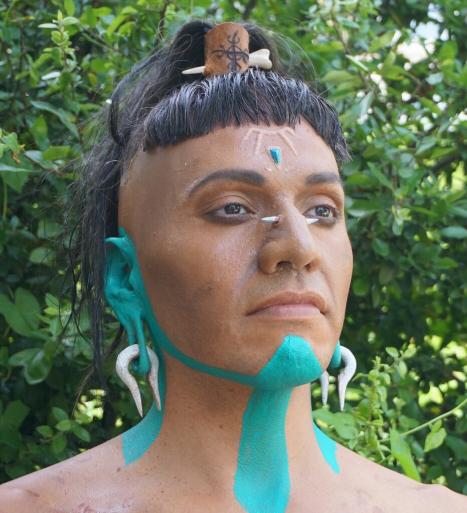 Das Bild zeigt einen Mann, der im Stil von Maya-Kriegern geschminkt wurde. Er hat am Nasenrücken ein Piercing mit einem Knochen. Er hat große Ohrlöcher mit Knochenschmuck. Auf der Stirn ist eine Skarifizierung angeschminkt. 